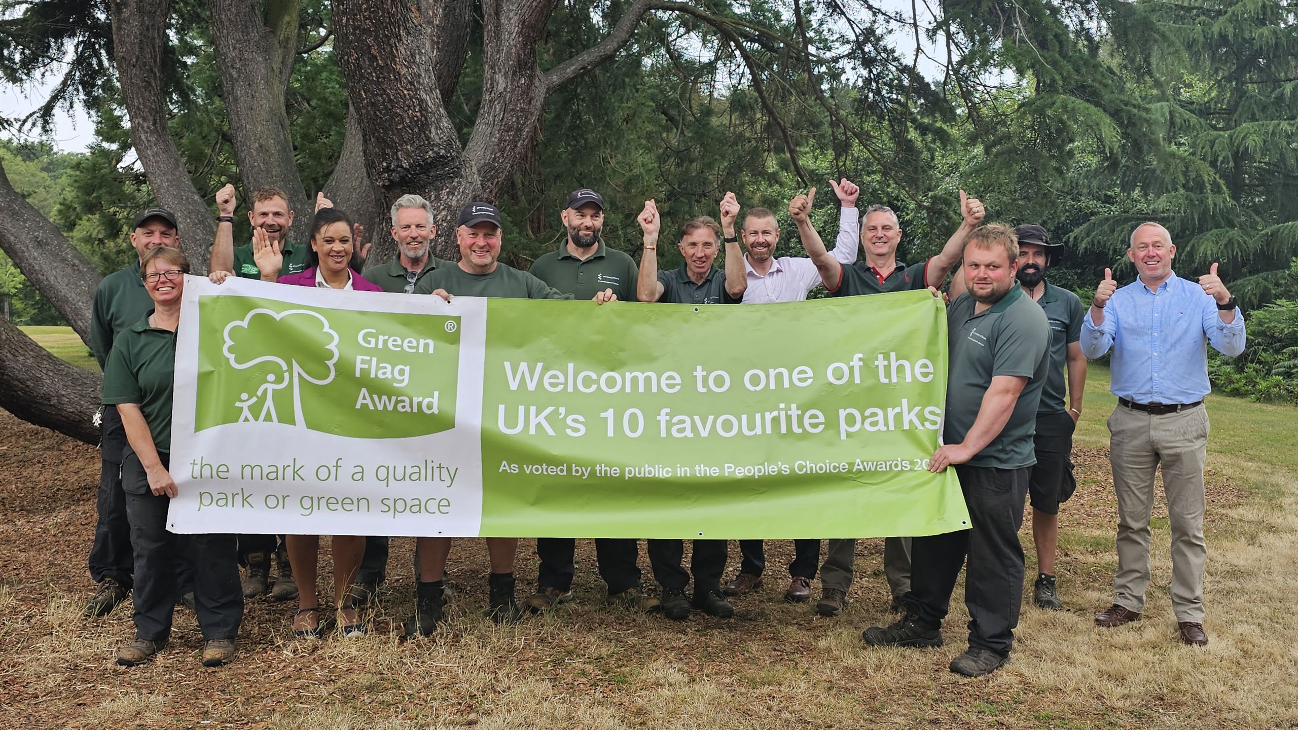 Staff from our Grounds team smile with their hands in the air. Two of them hold a banner which reads 'Green Flag Award, the mark of a quality park or green space. Welcome to one of the UK's 10 favourite parks as voted by the public in the People's Choice Awards 2023'.