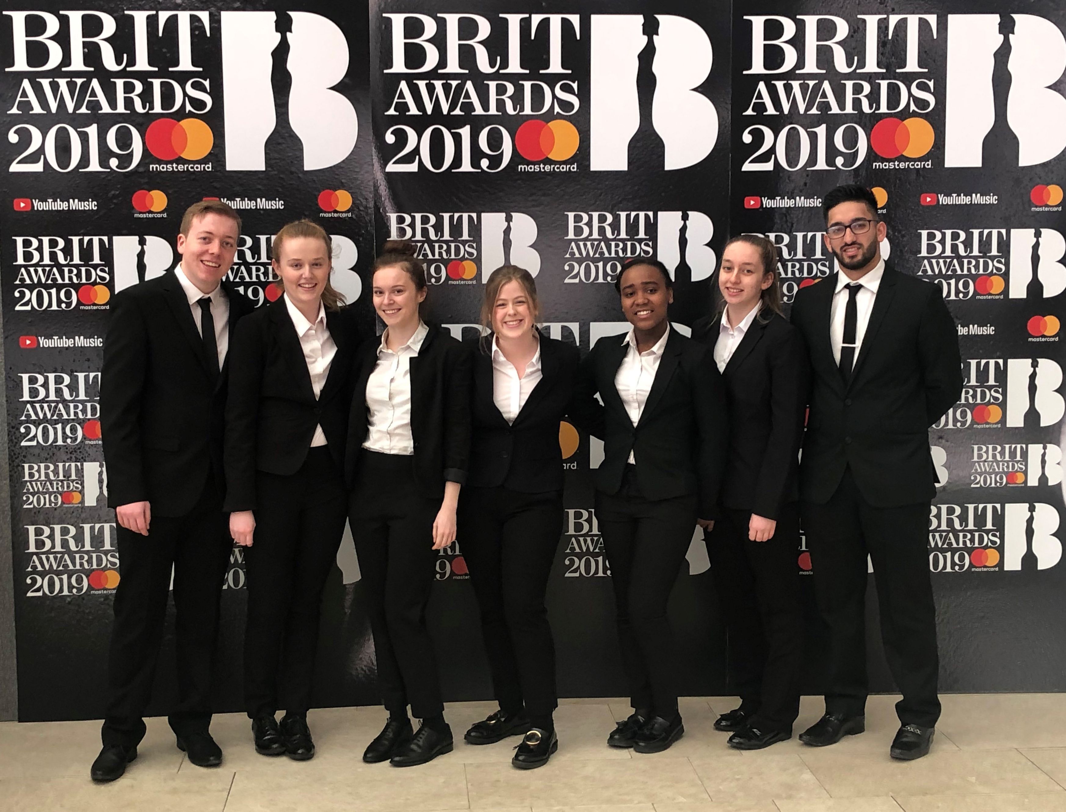 A group of smiling Edge Hotel School students, all wearing smart black suits and white shirts, line up in front of a BRIT Awards 2019 back drop