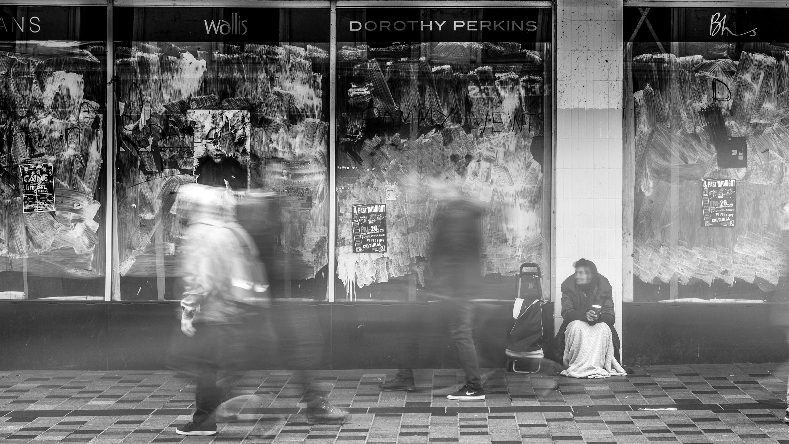 Black and white picture of a homeless lady sat in front of a series of boarded up shops