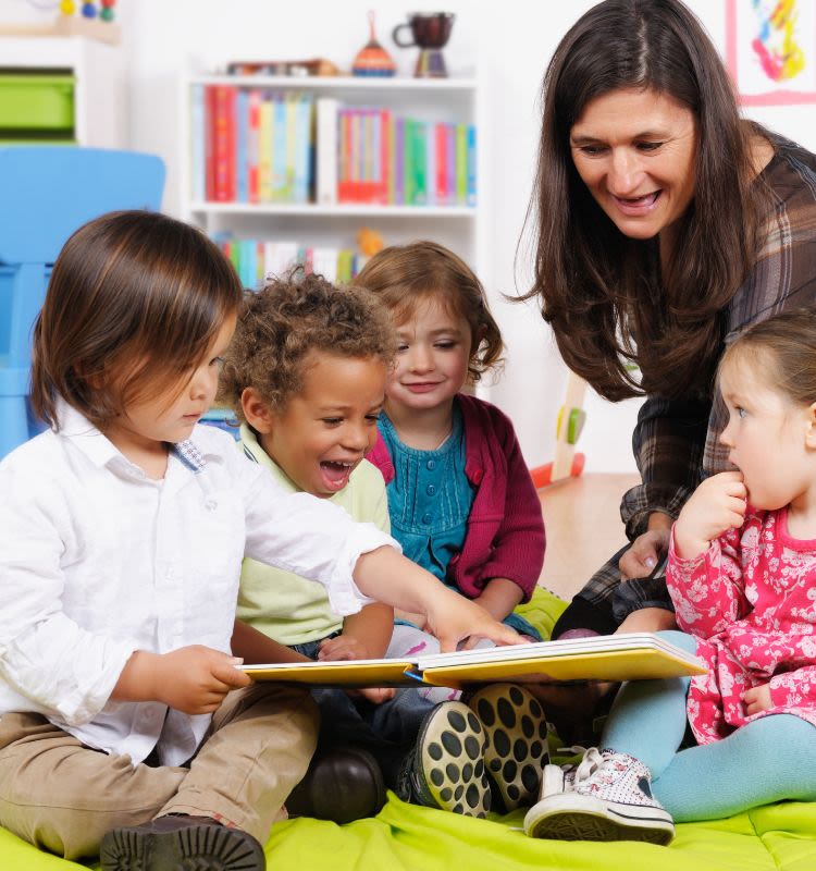 A woman talks with a group of toddlers as they look at a book together in a nursery
