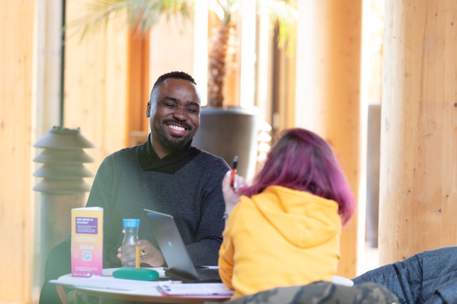 Two students laugh at a table in a sunny foyer of Essex Business School.